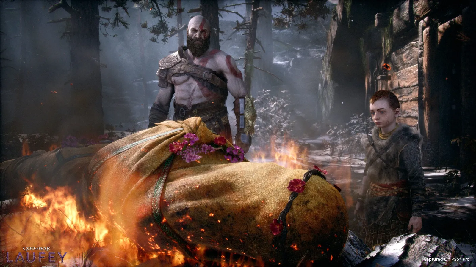 Kratos and a young Atreus stand solemnly beside a flower-covered funeral pyre holding a shrouded body in a dark snowy forest